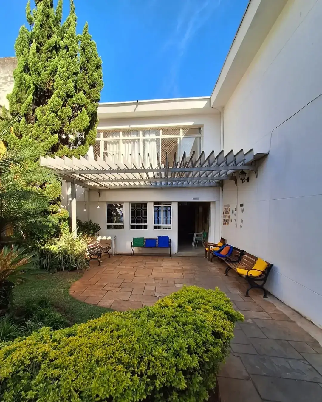 Fachada do casarão da Mental Sênior, com jardim, pergolado e bancos amarelos sob o céu azul