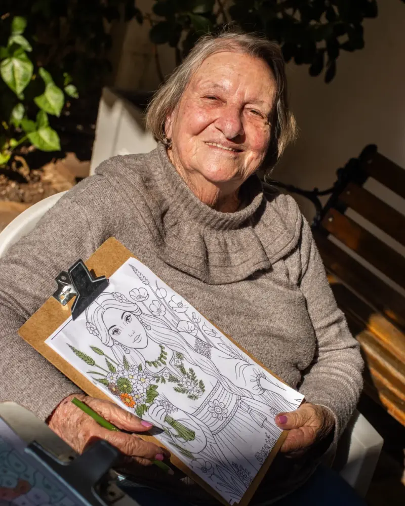 Residente sorridente segura um desenho que está colorindo, ao sol, durante atividade cognitiva